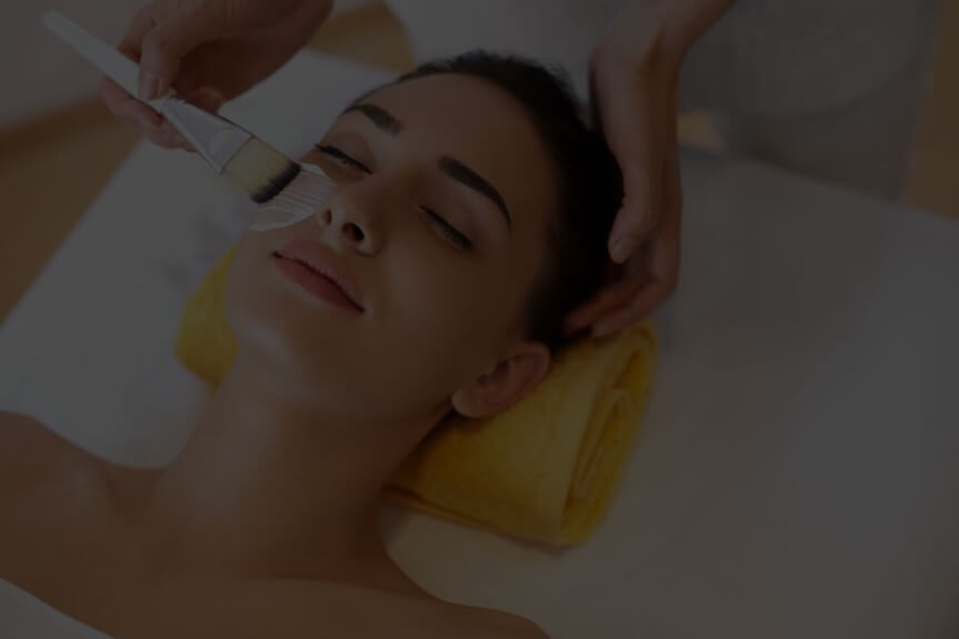 Person lying down with eyes closed, receiving a facial treatment with a brush applying cream.